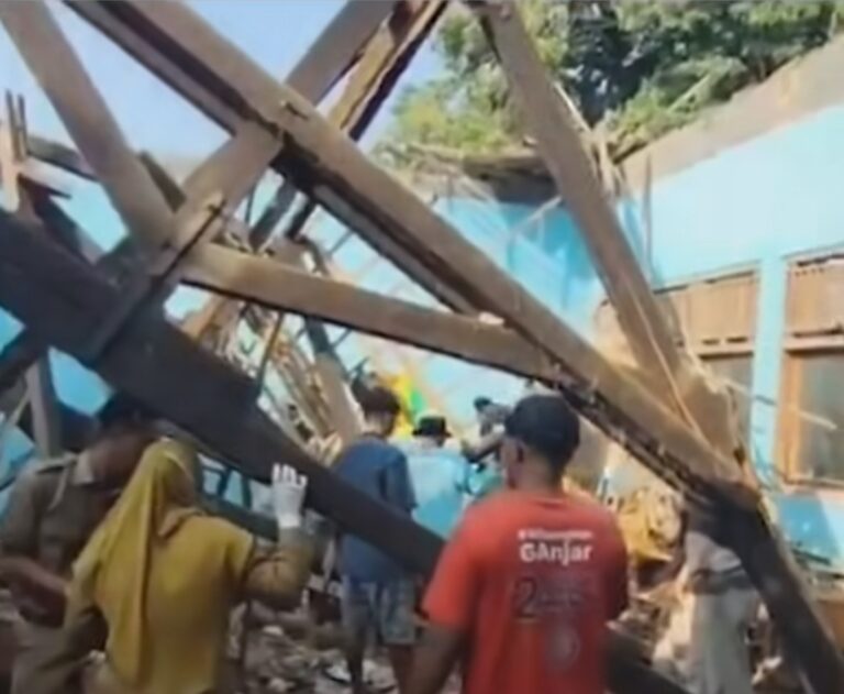People stand amid a tangle of large wooden beams and debris from a collapsed construction structure, with a blue tarpaulin in the background and onlookers nearby.