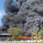 Large plume of thick black smoke and orange flames rising behind a white fence along a roadside—fire visible in daytime sky.