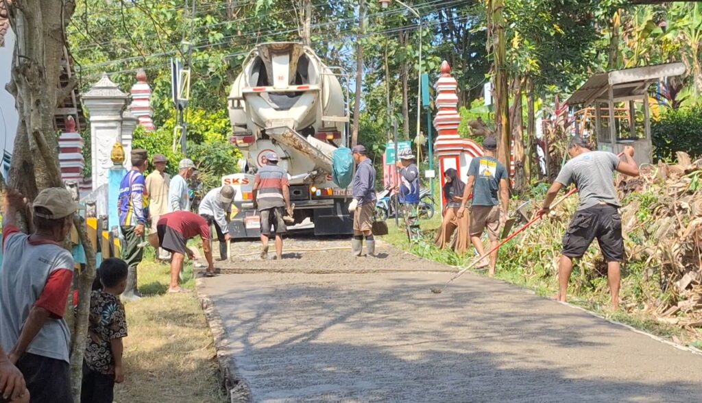 Workers spread and level fresh concrete on a road beside a cement mixer truck while onlookers watch from both sides.