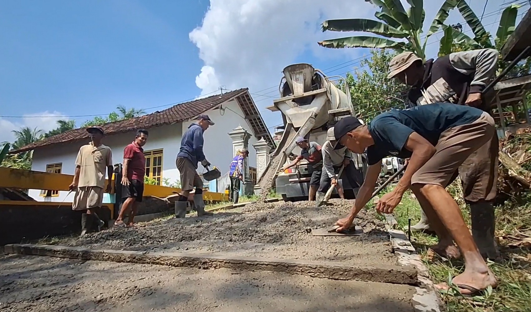 Group of workers repaving a small road in a rural neighborhood, using a concrete mixer and shovels while a few men help spread gravel and level the surface.