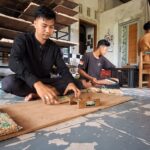 Three men sit on the floor in a workshop, packing snack bags into cardboard sheets.