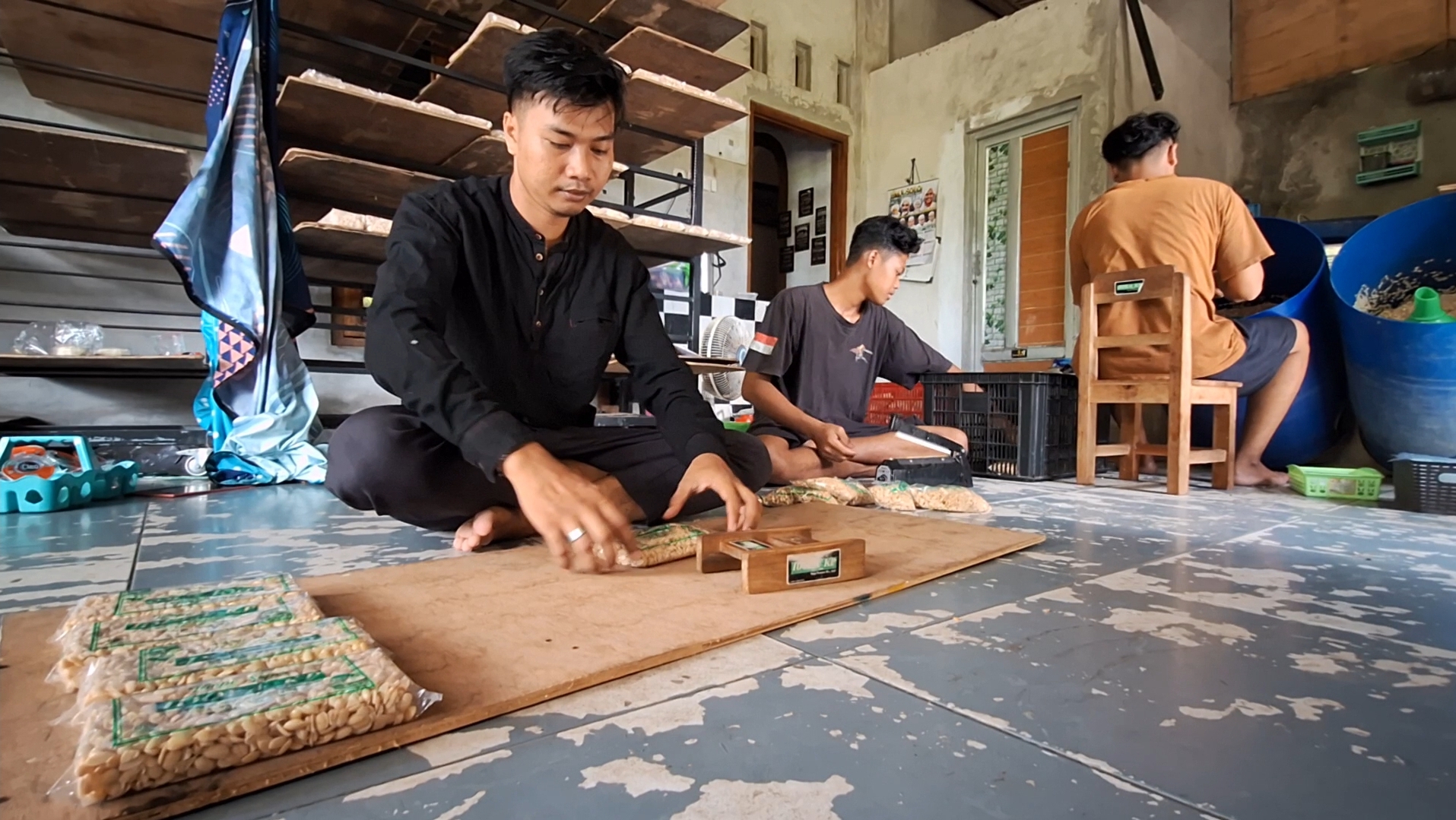 Three men sit on the floor in a workshop, packing snack bags into cardboard sheets.
