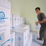 Man standing on a stack of Zamzam Water boxes, placing another box in a storage room.