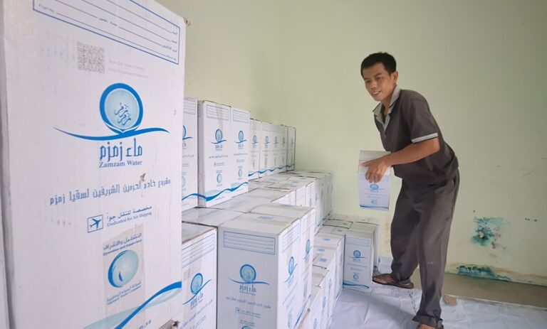 Man standing on a stack of Zamzam Water boxes, placing another box in a storage room.