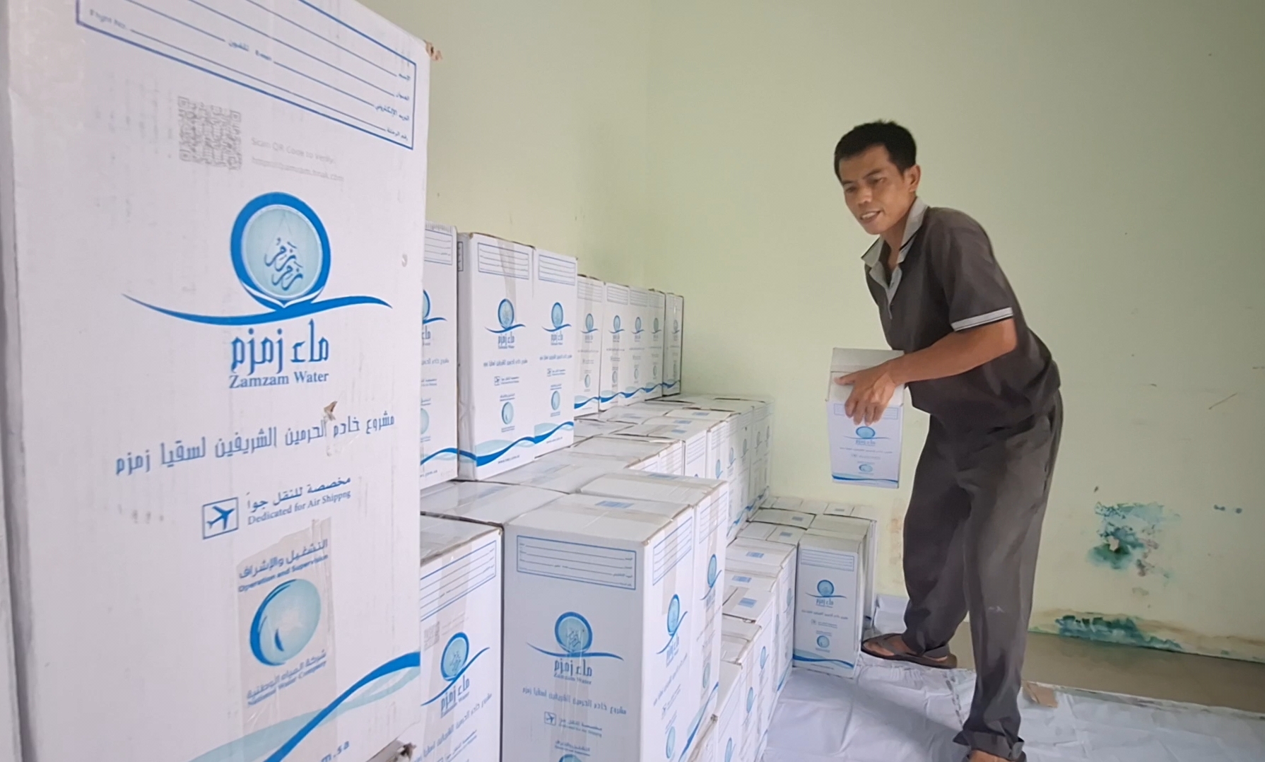 Man standing on a stack of Zamzam Water boxes, placing another box in a storage room.