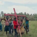 Group of men carrying a large red cross through a grassy rural field, some wearing grass skirts, with palm trees in the background.
