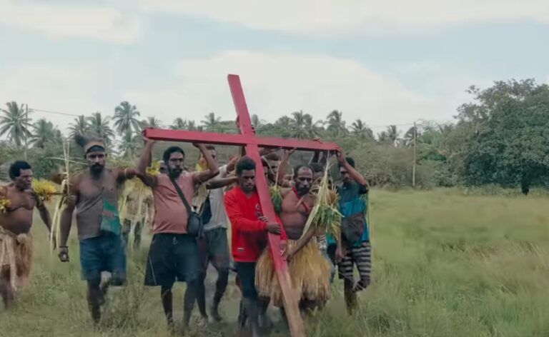 Screenshot_20260520_151720_YouTube - Mabur.co Group of men carrying a large red cross through a grassy rural field, some wearing grass skirts, with palm trees in the background.