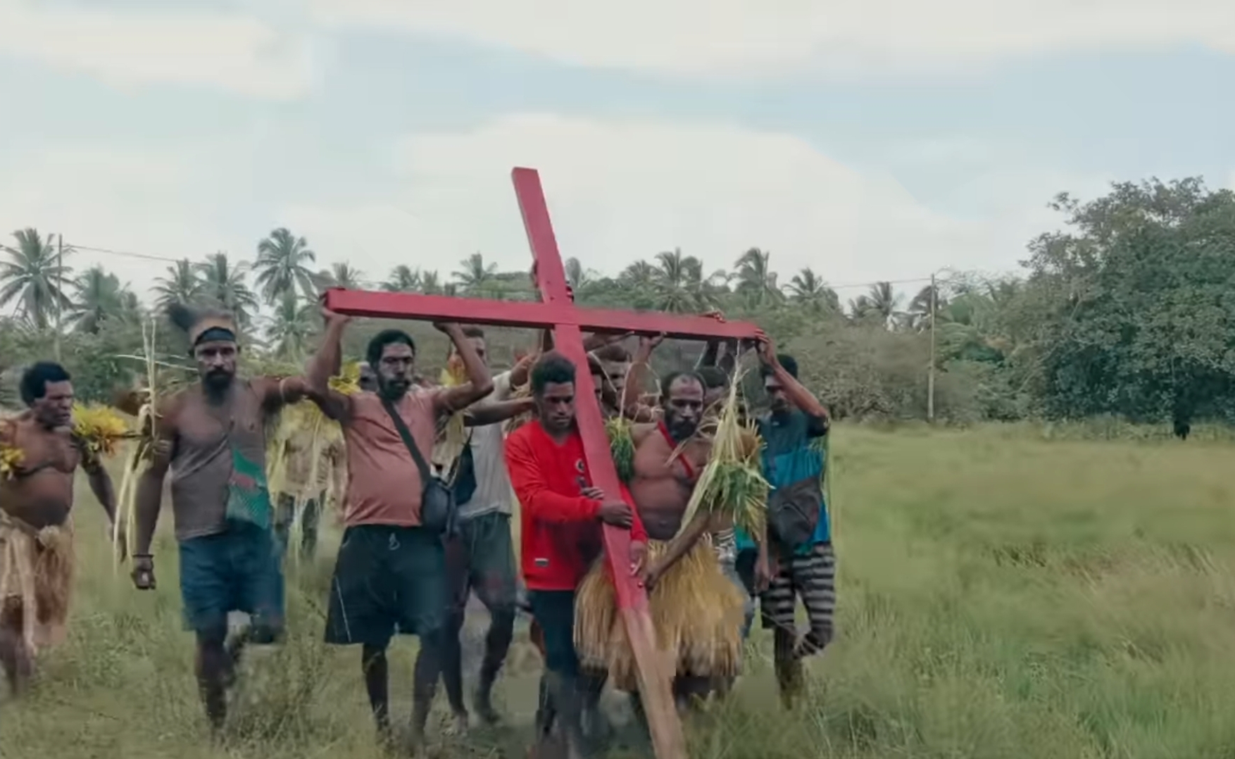 Group of men carrying a large red cross through a grassy rural field, some wearing grass skirts, with palm trees in the background.