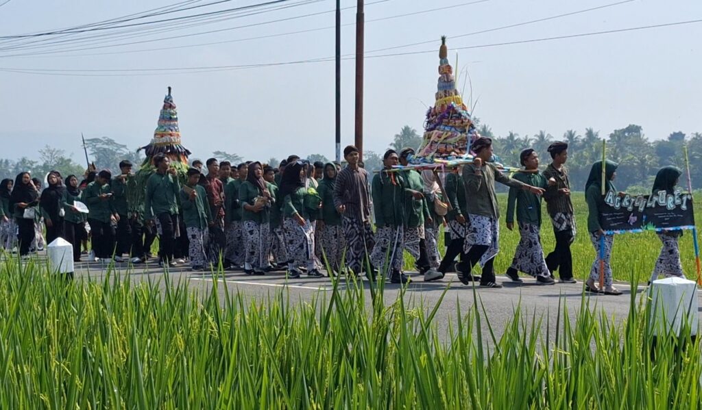 Screenshot_20260520_185848_Gallery - Mabur.co Group of people in green shirts marching along a road, carrying a tall, colorful ceremonial tower in a festival procession, with rice fields in the foreground.