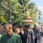 Parade with participants in green striped outfits walking along a palm-lined street, a colorful layered fruit tower behind them.