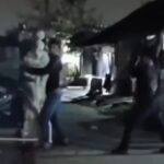 Two people appear to be in a street fight at night, one in light clothing and the other in dark clothing near a doorway.