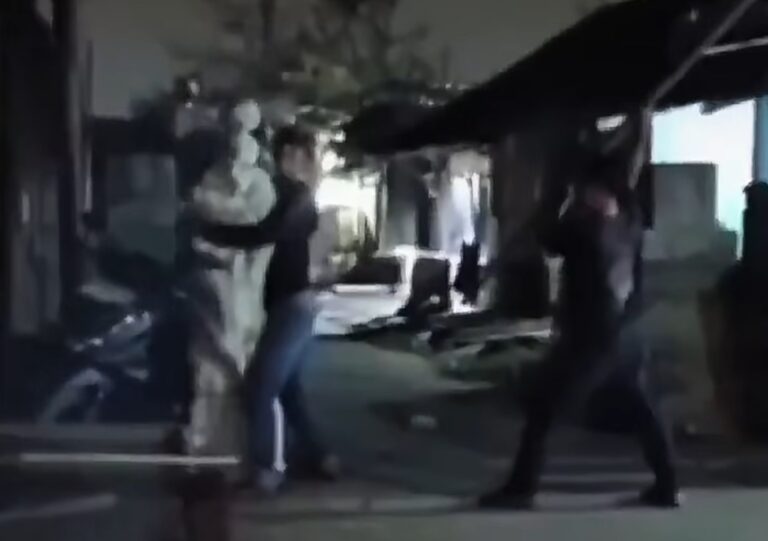 Two people appear to be in a street fight at night, one in light clothing and the other in dark clothing near a doorway.