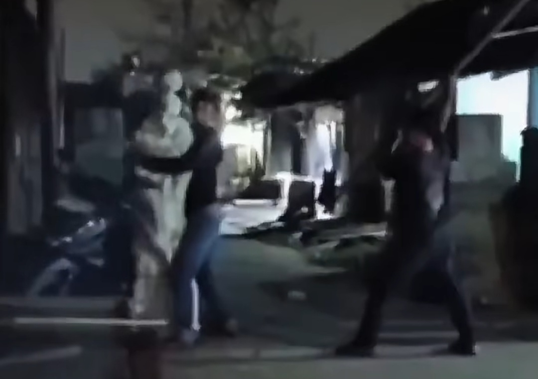 Two people appear to be in a street fight at night, one in light clothing and the other in dark clothing near a doorway.