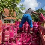 A worker in a striped shirt hands a pink gas cylinder to another person on a truck loaded with many pink LPG cylinders outdoors.