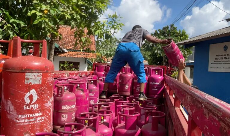 Screenshot_20260522_172833_Gallery - Mabur.co A worker in a striped shirt hands a pink gas cylinder to another person on a truck loaded with many pink LPG cylinders outdoors.