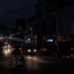 Nighttime city street with car headlights and a motorcycle on a dimly lit road, storefronts along the block glow in the distance.