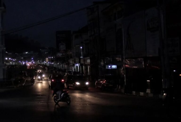 Nighttime city street with car headlights and a motorcycle on a dimly lit road, storefronts along the block glow in the distance.