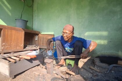 Older man in a blue shirt and brown cap sits cross‑legged in a workshop, handling wooden tools near a workbench against a green wall.
