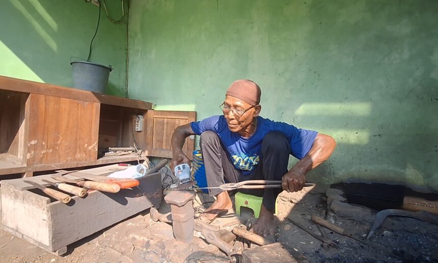 Screenshot_20260525_104805_Gallery - Mabur.co Older man in a blue shirt and brown cap sits cross‑legged in a workshop, handling wooden tools near a workbench against a green wall.