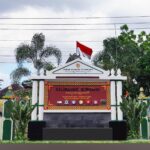 Ornate entrance gate with a red sign panel, Indonesian flag on top, and tropical plants flanking white columns; banners on both sides indicate a village office.