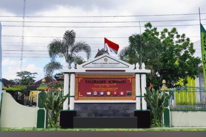 Ornate entrance gate with a red sign panel, Indonesian flag on top, and tropical plants flanking white columns; banners on both sides indicate a village office.