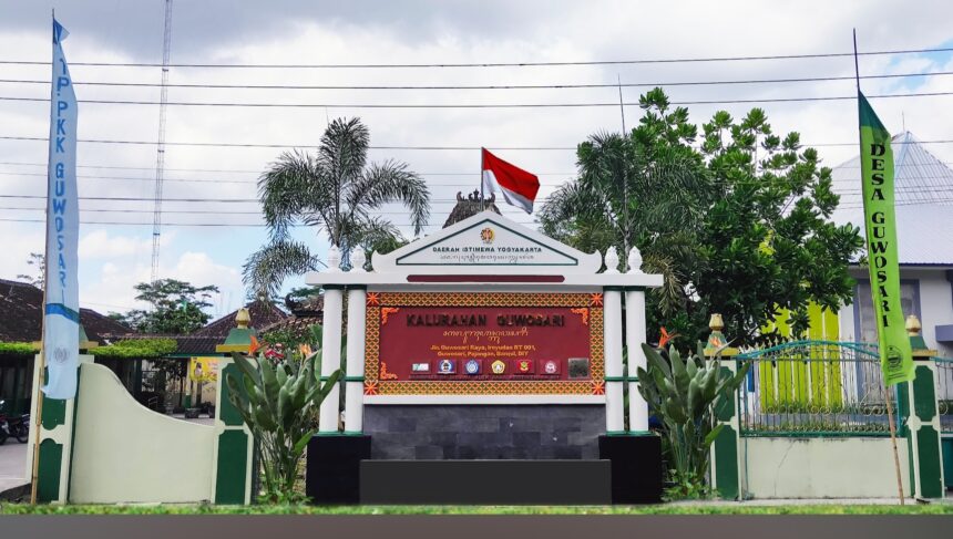 Ornate entrance gate with a red sign panel, Indonesian flag on top, and tropical plants flanking white columns; banners on both sides indicate a village office.