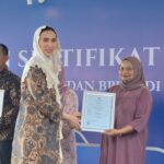 Award ceremony on stage: woman in a patterned dress hands a framed certificate to a woman in a mauve hijab; two men stand with certificates, blue backdrop behind them.