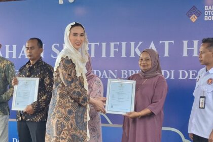 Award ceremony on stage: woman in a patterned dress hands a framed certificate to a woman in a mauve hijab; two men stand with certificates, blue backdrop behind them.