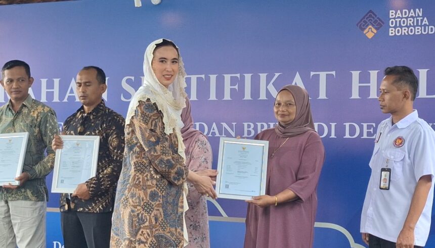 Award ceremony on stage: woman in a patterned dress hands a framed certificate to a woman in a mauve hijab; two men stand with certificates, blue backdrop behind them.