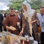 A woman in a patterned dress and white lace hijab points at a table of snacks while a man beside her holds a cup, surrounded by onlookers at an outdoor event.