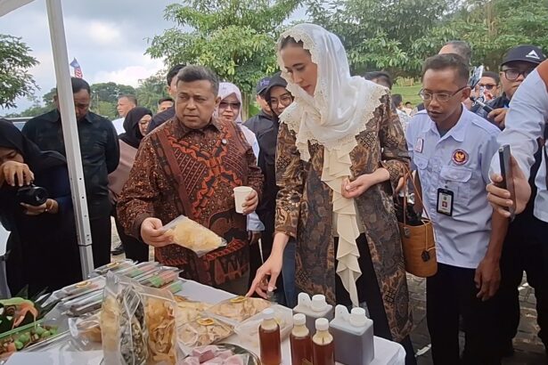 A woman in a patterned dress and white lace hijab points at a table of snacks while a man beside her holds a cup, surrounded by onlookers at an outdoor event.