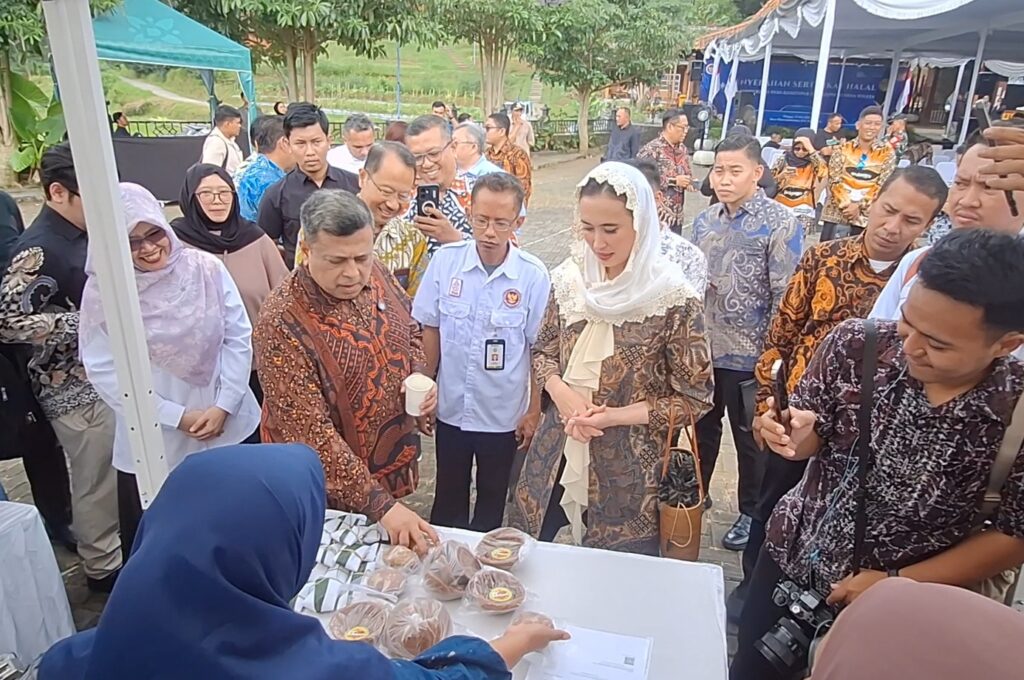 Diverse group of men and women in batik and hijabs gather around an outdoor table displaying wrapped pastries, with a vendor in a blue headscarf arranging items.
