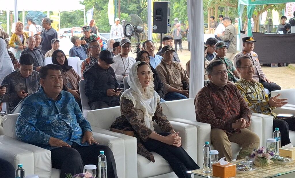 D group of formally dressed attendees seated on white sofas at an outdoor ceremony under a canopy, with a table of water bottles in front.