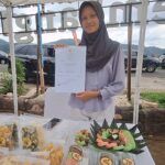 Woman in hijab displaying a certificate at an outdoor market booth with snacks on the table.",