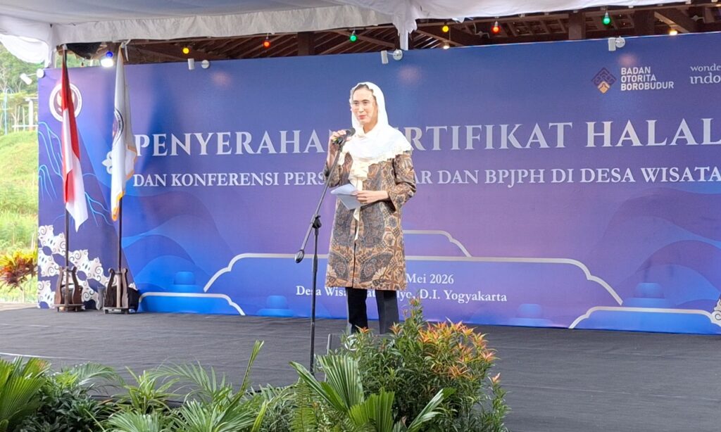 Woman in a patterned dress and white headscarf speaking into a microphone on a blue event stage with flags and plants in the foreground.