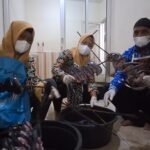 Three people wearing masks and gloves sit on the floor around large buckets, cutting or sorting seafood; two women in yellow headscarves and a man in a dark cap participate in a processing activity in an indoor setting.