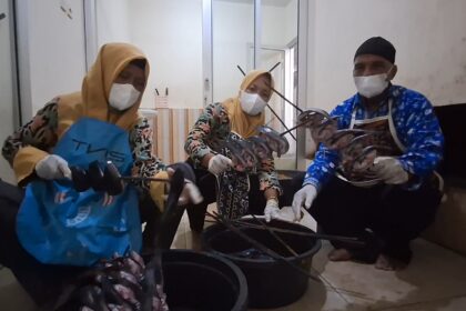 Three people wearing masks and gloves sit on the floor around large buckets, cutting or sorting seafood; two women in yellow headscarves and a man in a dark cap participate in a processing activity in an indoor setting.