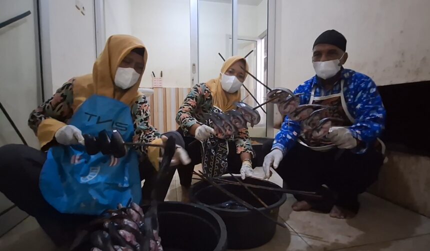 Three people wearing masks and gloves sit on the floor around large buckets, cutting or sorting seafood; two women in yellow headscarves and a man in a dark cap participate in a processing activity in an indoor setting.