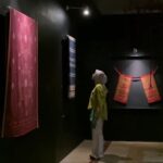 Dim museum gallery with a large red patterned textile hanging on the left and a blue striped fabric toward the back; a person wearing a headscarf and yellow top stands nearby, viewing the displays.