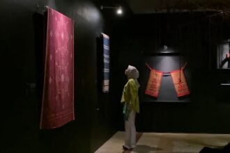 Dim museum gallery with a large red patterned textile hanging on the left and a blue striped fabric toward the back; a person wearing a headscarf and yellow top stands nearby, viewing the displays.