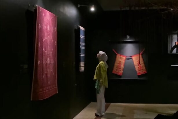 Dim museum gallery with a large red patterned textile hanging on the left and a blue striped fabric toward the back; a person wearing a headscarf and yellow top stands nearby, viewing the displays.