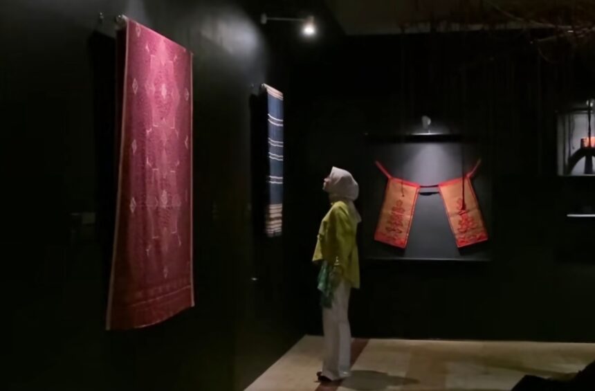 Dim museum gallery with a large red patterned textile hanging on the left and a blue striped fabric toward the back; a person wearing a headscarf and yellow top stands nearby, viewing the displays.