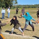 Children in colorful modest clothing run on a sunny grassy field, playing a chase game as adults watch in the background with goalposts nearby.