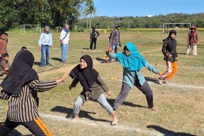 Children in colorful modest clothing run on a sunny grassy field, playing a chase game as adults watch in the background with goalposts nearby.