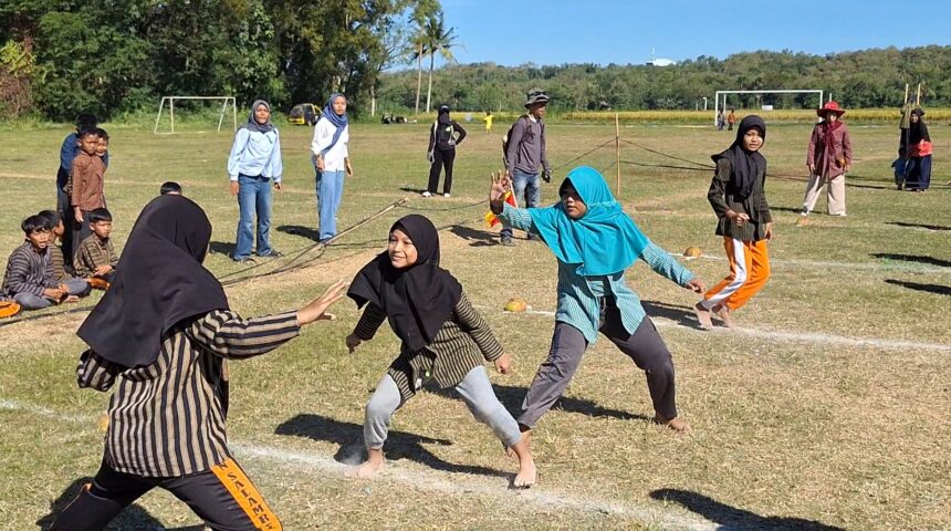 Children in colorful modest clothing run on a sunny grassy field, playing a chase game as adults watch in the background with goalposts nearby.