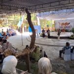 Outdoor festival scene with a circular stage under a canopy and a banner; audience seated around, many women in blue hijabs. There are speakers on stage and presenters.