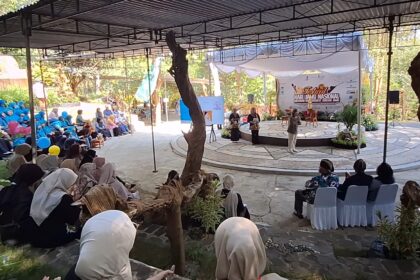 Outdoor festival scene with a circular stage under a canopy and a banner; audience seated around, many women in blue hijabs. There are speakers on stage and presenters.