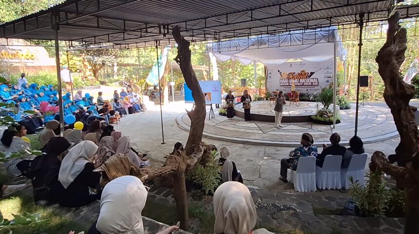 Outdoor festival scene with a circular stage under a canopy and a banner; audience seated around, many women in blue hijabs. There are speakers on stage and presenters.