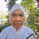 Close-up of an elderly man in a white turban outdoors, with greenery in the background and a smartphone visible in the foreground.