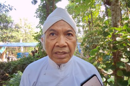 Close-up of an elderly man in a white turban outdoors, with greenery in the background and a smartphone visible in the foreground.